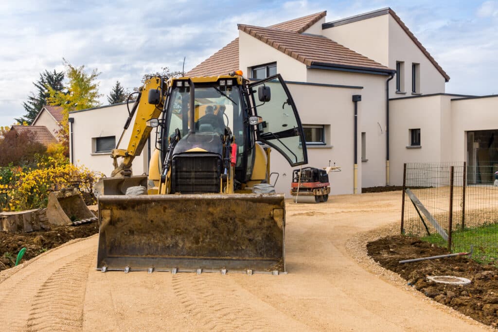 Travaux de construction : engins et nouvelle maison Chargeuse-pelleteuse jaune devant une maison moderne en construction, travail de terrassement sur chemin de gravier en automne.