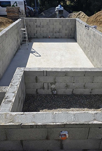 Construction piscine : Murs en parpaings et dalle béton coulée. Bassin de piscine en construction (parpaings/dalle béton). Une marche ou zone technique en gravier est visible. Matériel de chantier en arrière-plan.