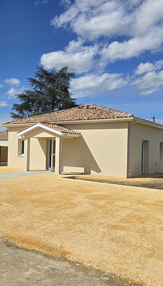 Maison neuve de plain-pied : façade beige et ciel bleu Plain-pied neuf à façade beige, toit en tuiles et porche d'entrée. Le jardin est couvert de gravier sous un grand cèdre et ciel bleu.
